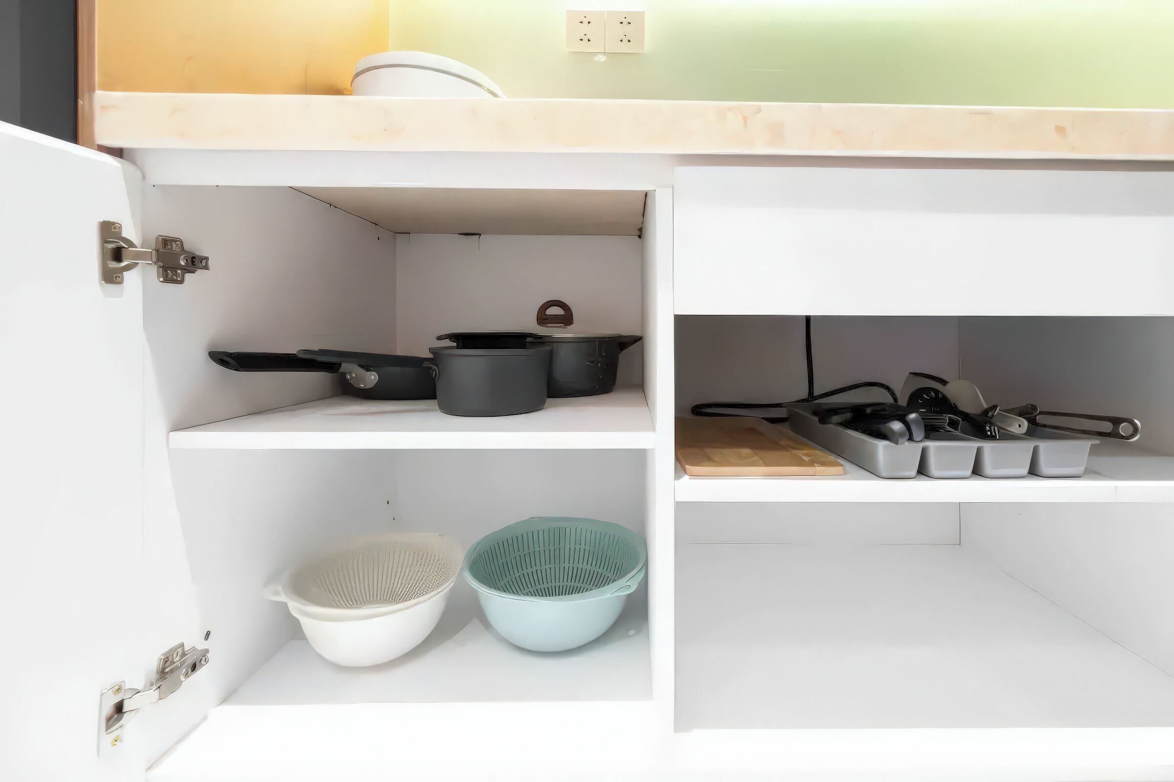 Una cocina con armarios blancos, una encimera y un lavabo. Hay ollas y sartenes sobre la encimera, un colador y una tabla de cortar.