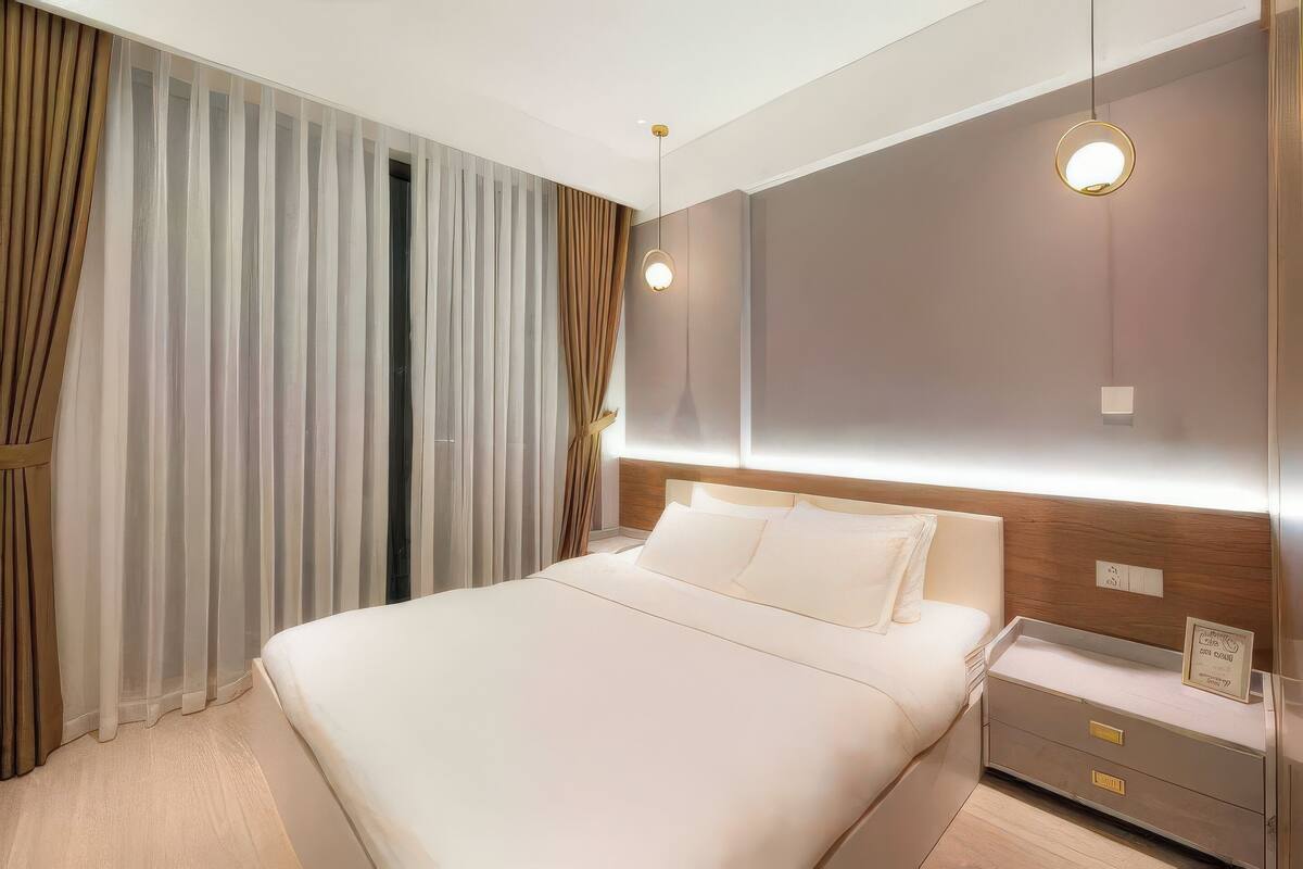 Habitació d'hotel moderna amb un llit gran, una tauleta de nit amb una làmpada i cortines translúcides.