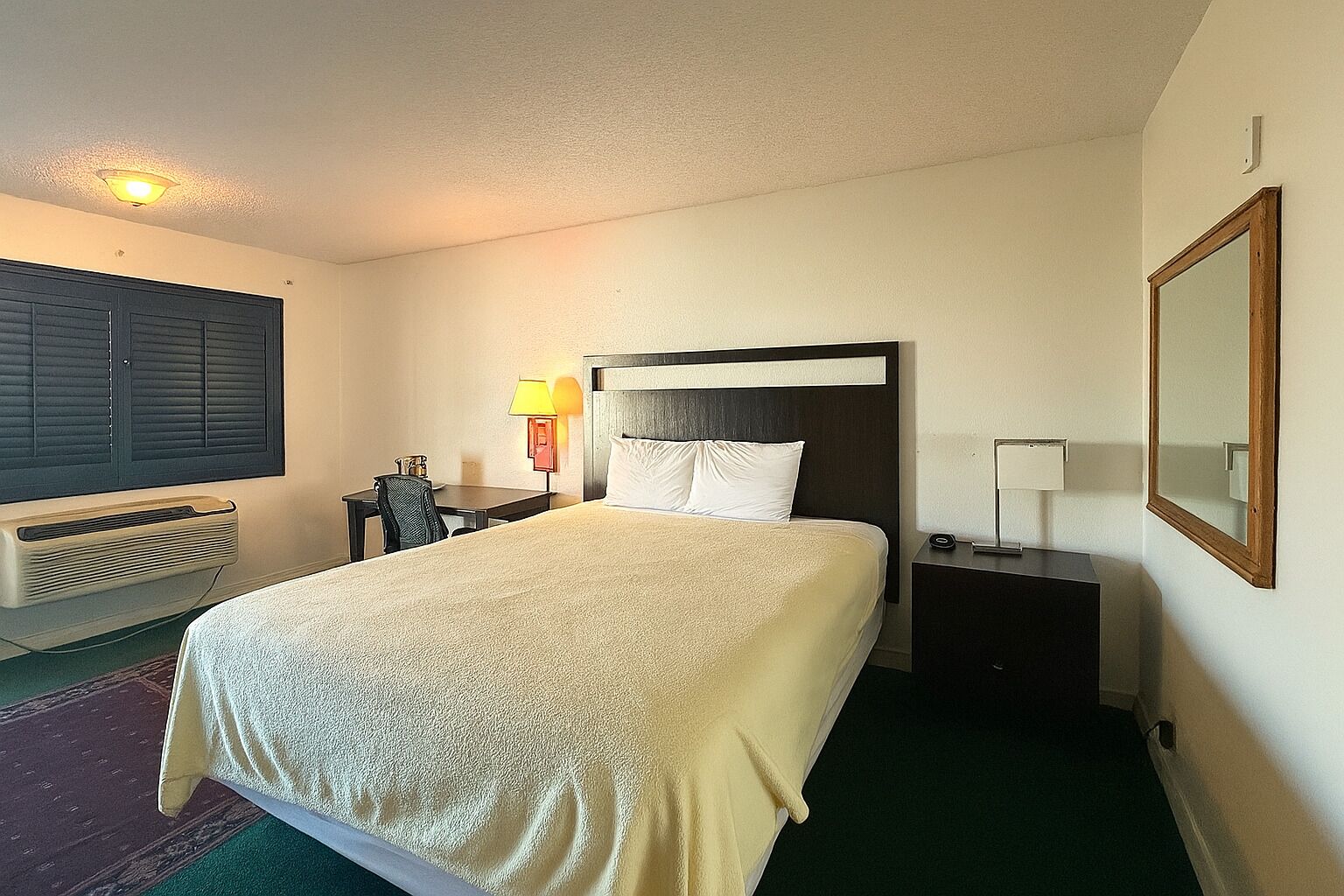 A hotel room with a bed, a desk, a chair, a lamp, a heater, and a mirror.