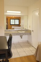 Un baño de hotel con lavabo, espejo y vistas a un dormitorio con cama y ventana.