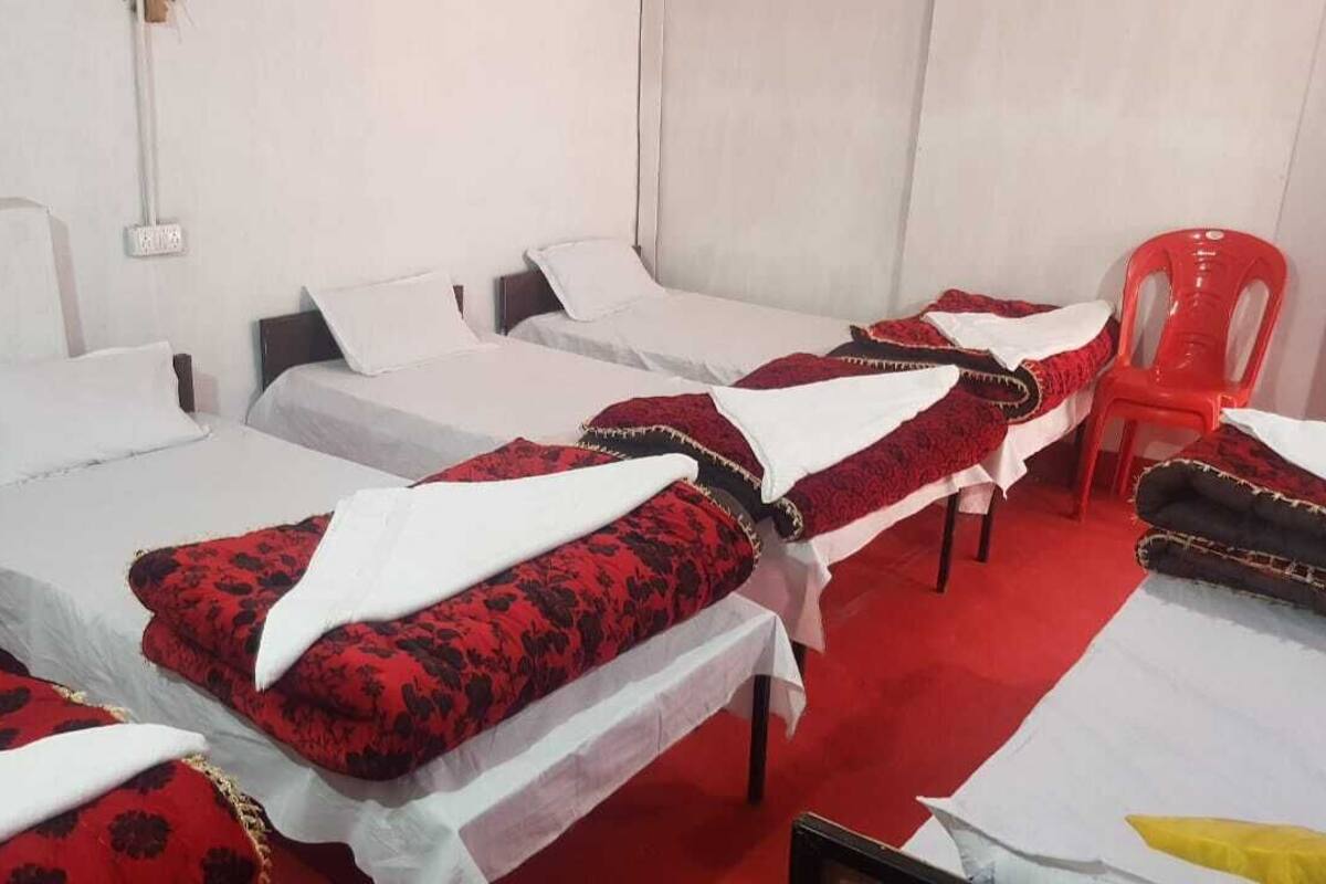 A hotel room with four single beds, each with a red and black patterned bedspread and white pillows. There is a red plastic chair and a white table in the room.