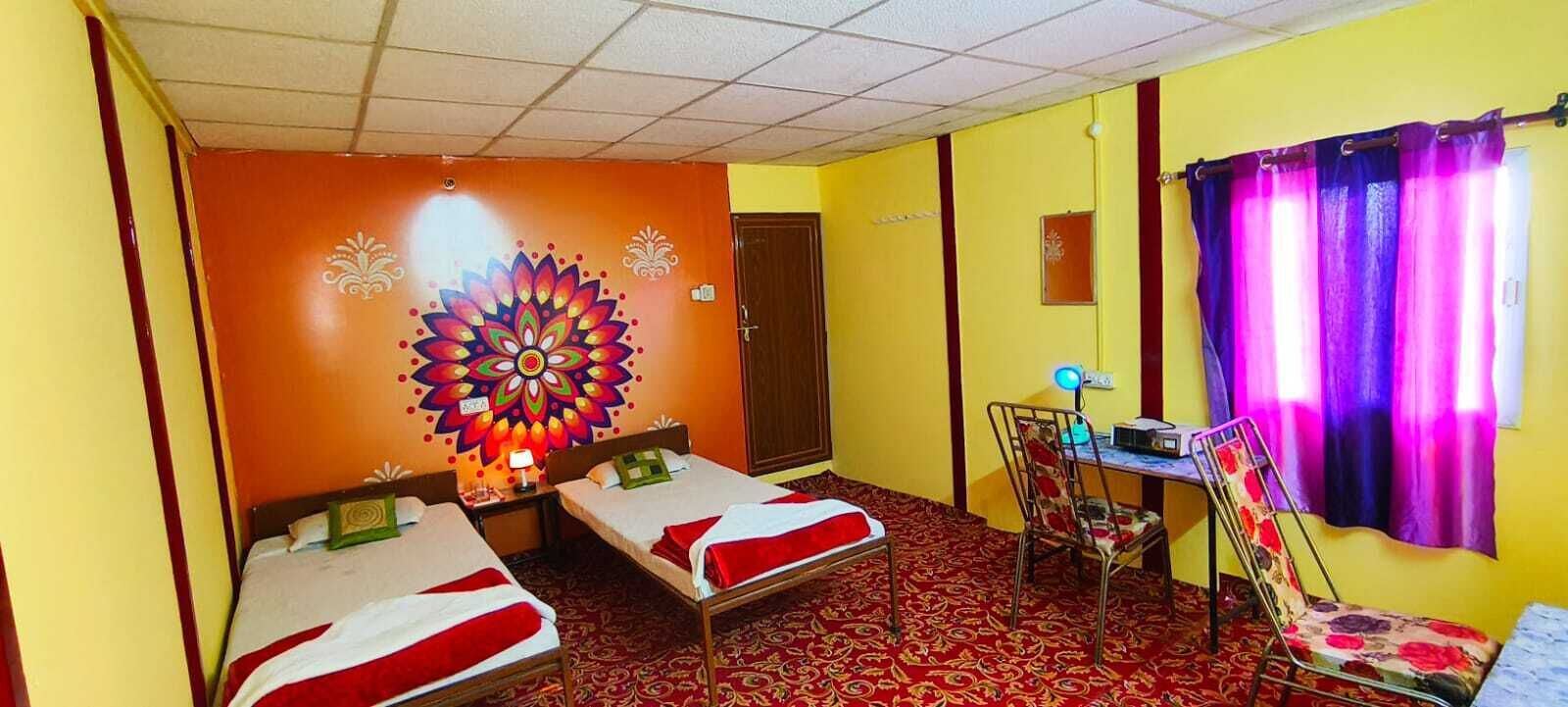 A room with two single beds, a desk with a chair, a colorful wall mural, and a patterned carpet.