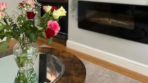A living room with a TV displaying a scenic landscape, a vase with flowers on a glass table, and a fireplace below. - Beautiful 4 Bed House in Sheffield (Sheffield)