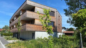 A modern multi-story building with a brick facade, balconies, and a garden area with various flowers.