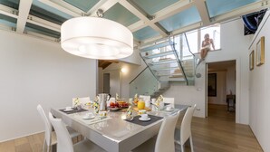Un espace repas moderne avec un escalier en verre, des meubles blancs et une table préparée pour le petit-déjeuner.