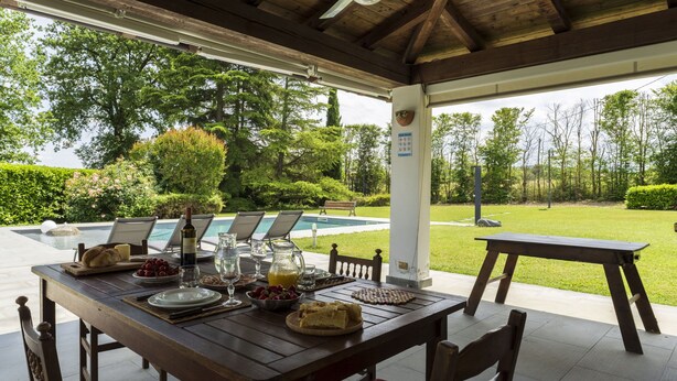 Un espace extérieur abrité, aménagé pour les repas, avec vue sur la piscine, comprenant une table en bois, des chaises et un banc.