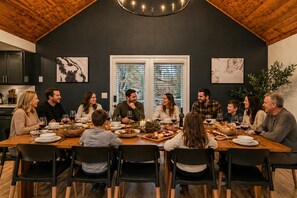 Una familia cenando en una cocina moderna con techos altos y vigas de madera, ventanas amplias y obras de arte en las paredes.