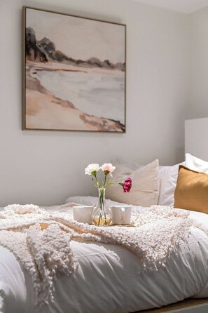 Uma cama bem arrumada com edredom branco e franjas decorativas, um vaso com flores e um quadro de uma cena de praia na parede.