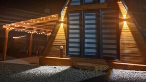 Une cabane en bois avec un toit triangulaire, de grandes fenêtres et une véranda couverte décorée de guirlandes lumineuses.