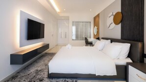 A modern bedroom with a large bed, a flat-screen TV mounted on the wall, a desk with a chair, and a window with blinds.