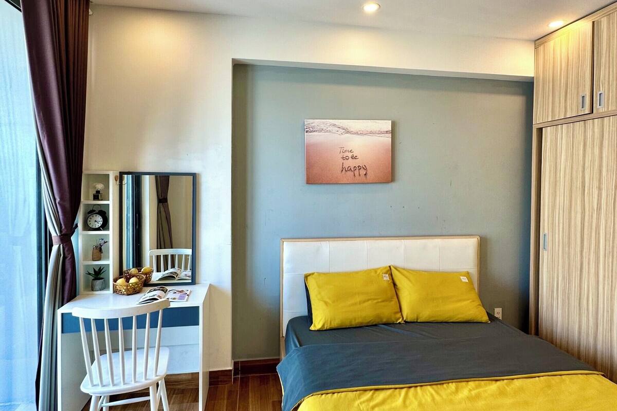A bedroom with a bed, a desk, and a chair. There is a painting on the wall and a window with curtains.
