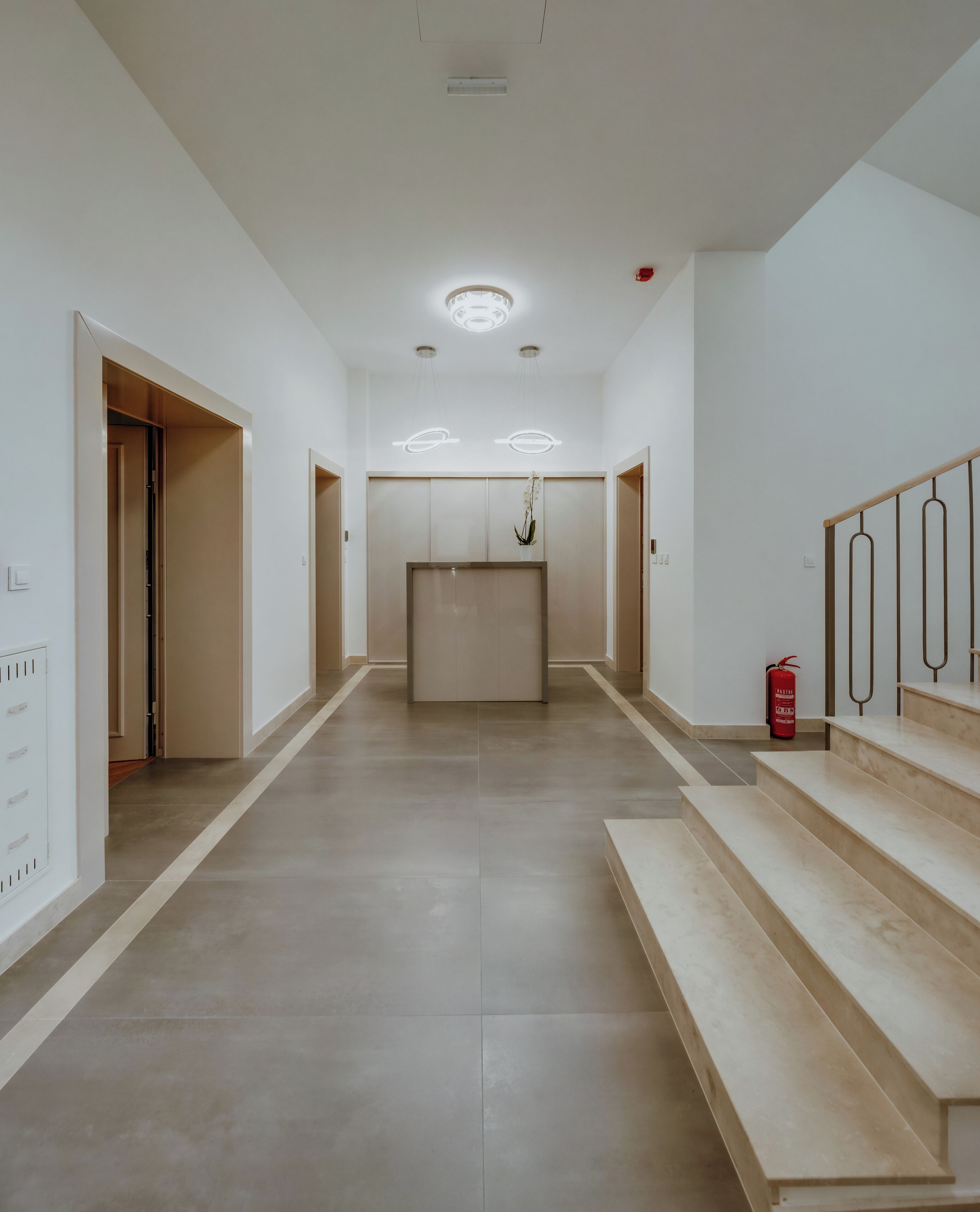 A spacious hallway with a central elevator, flanked by doors on both sides, and a fire extinguisher on the right.