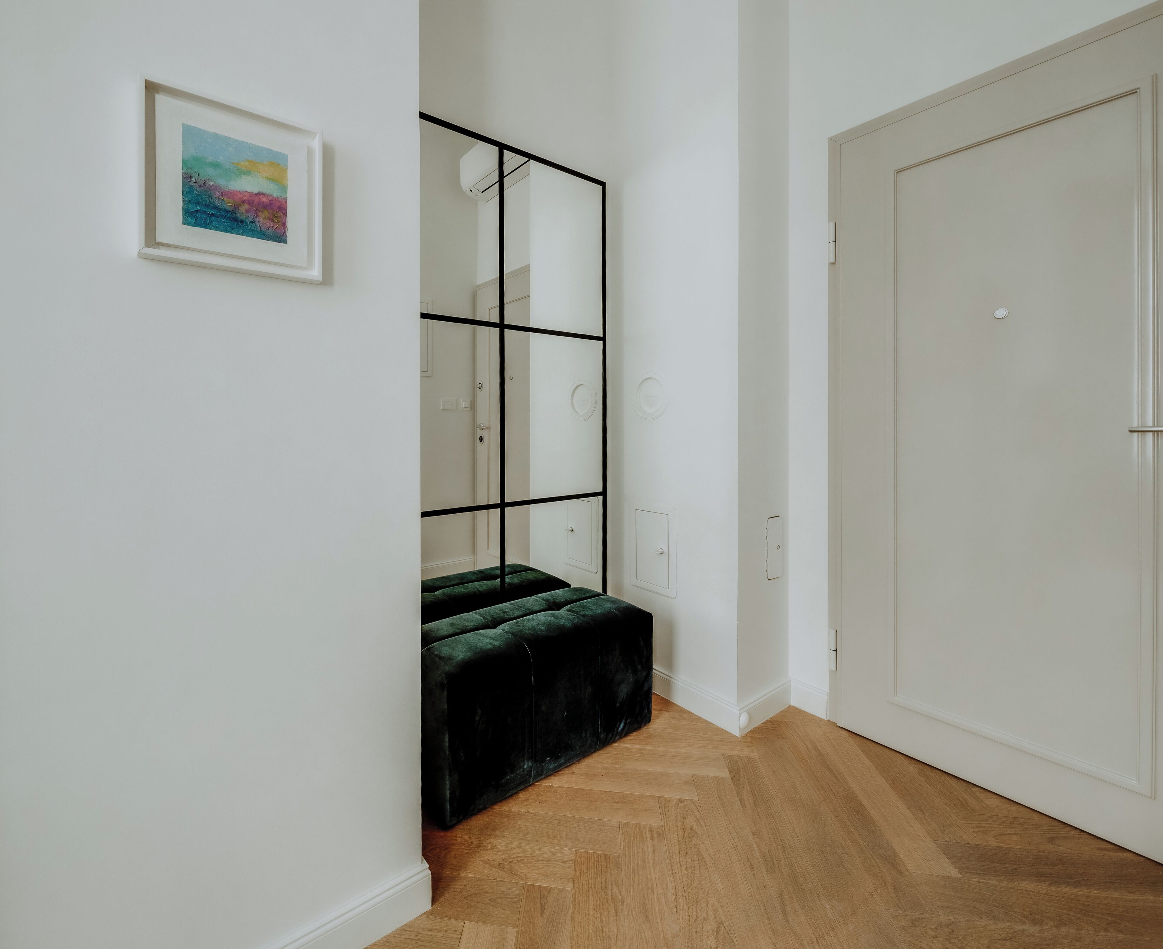 Een gang met een houten vloer, een spiegel met een zwart frame, een groene gestoffeerde bank en een witte deur.