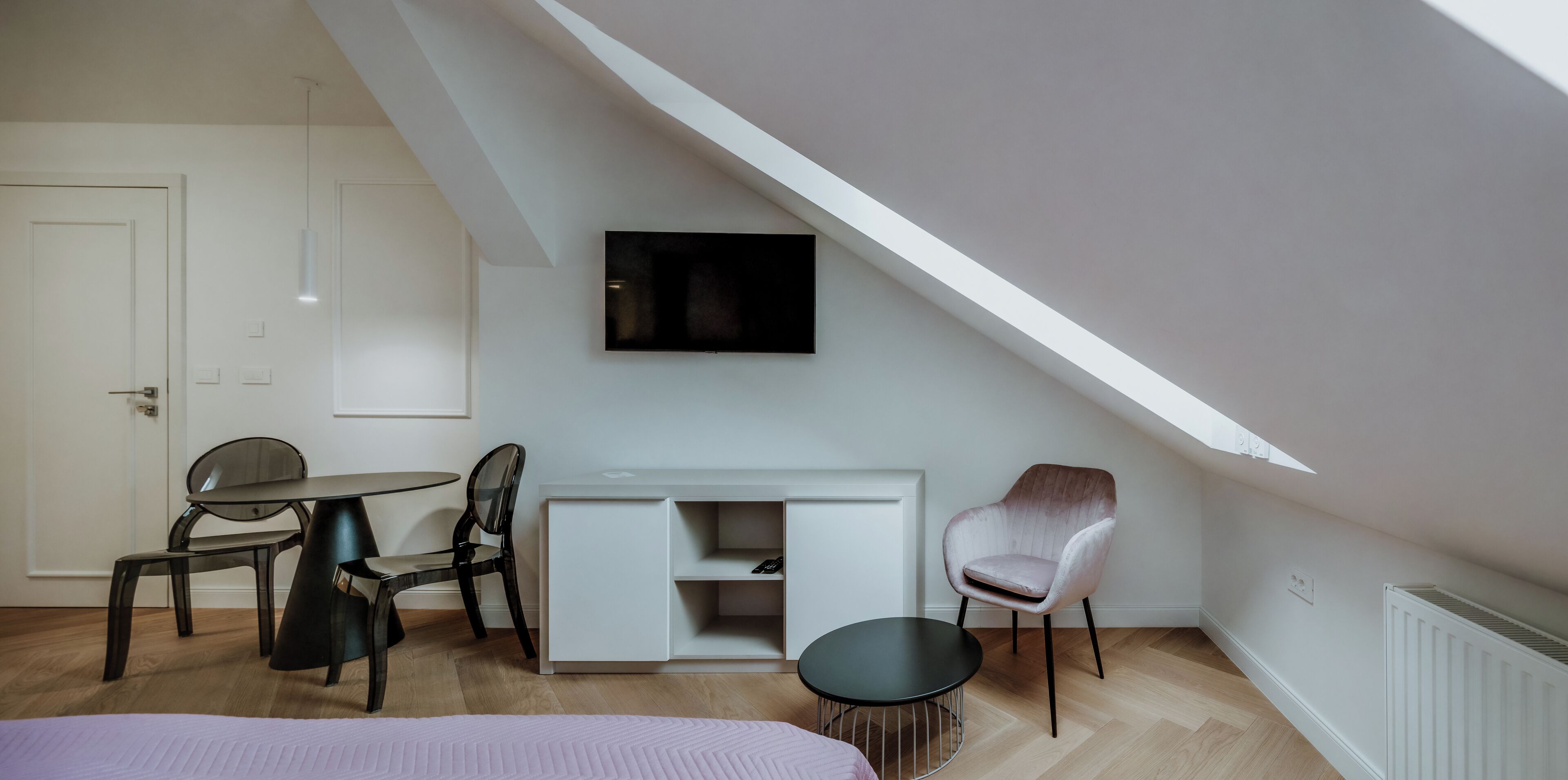 A modern, well-lit room with a loft-style ceiling, a white desk with a flat-screen TV, a comfortable chair, and a small round table.