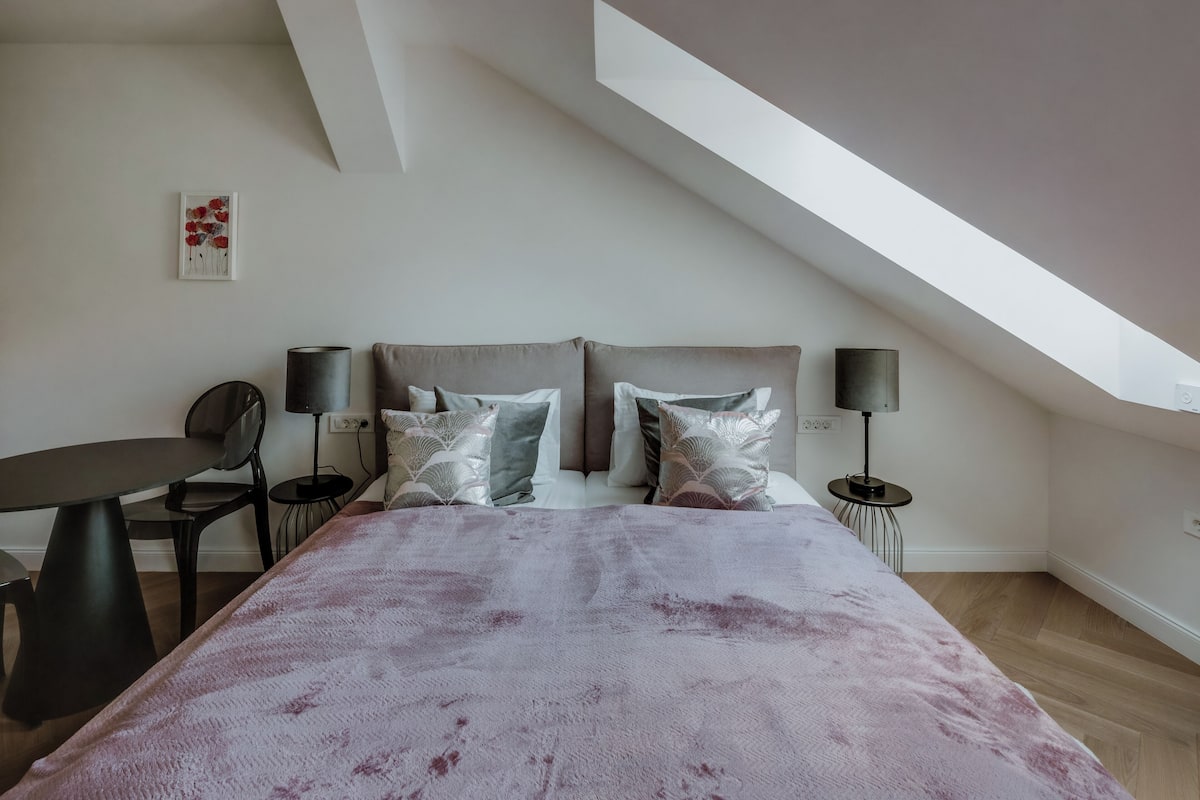 Une chambre avec un plafond en pente, un lit recouvert d’une couverture à motifs, deux lampes de chevet, une chaise et une table.