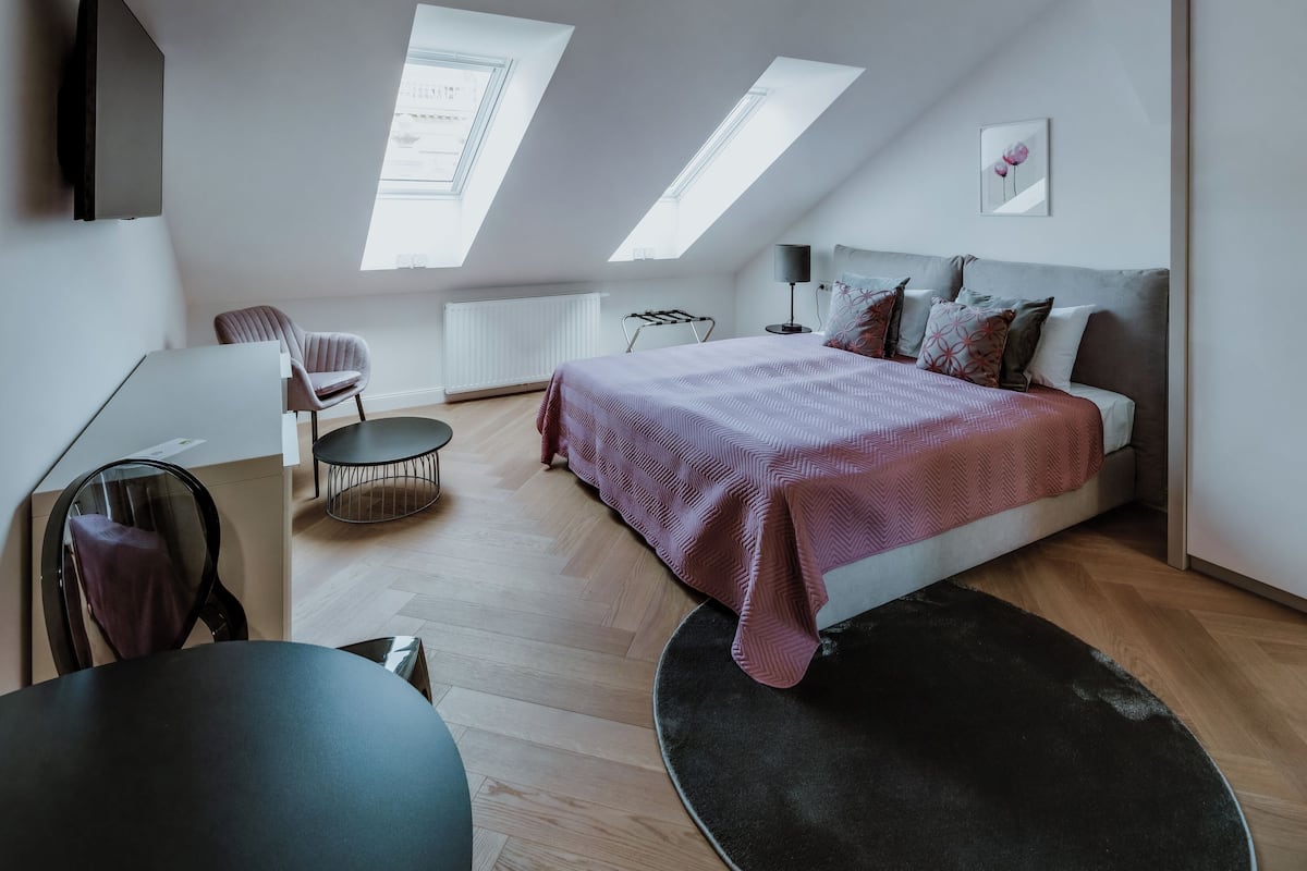 Une chambre moderne avec un plafond de style industriel, un lit recouvert d’une couverture rose, un bureau avec une chaise et un radiateur.