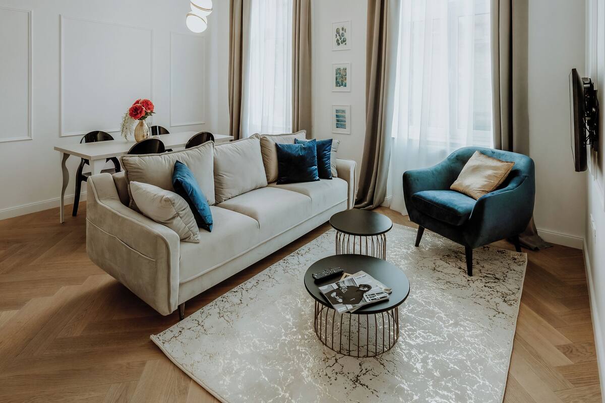 Un salon moderne avec un canapé, une table basse et un fauteuil bleu. On y trouve également une table à manger avec des chaises, un téléviseur fixé au mur et de grandes fenêtres avec des rideaux.