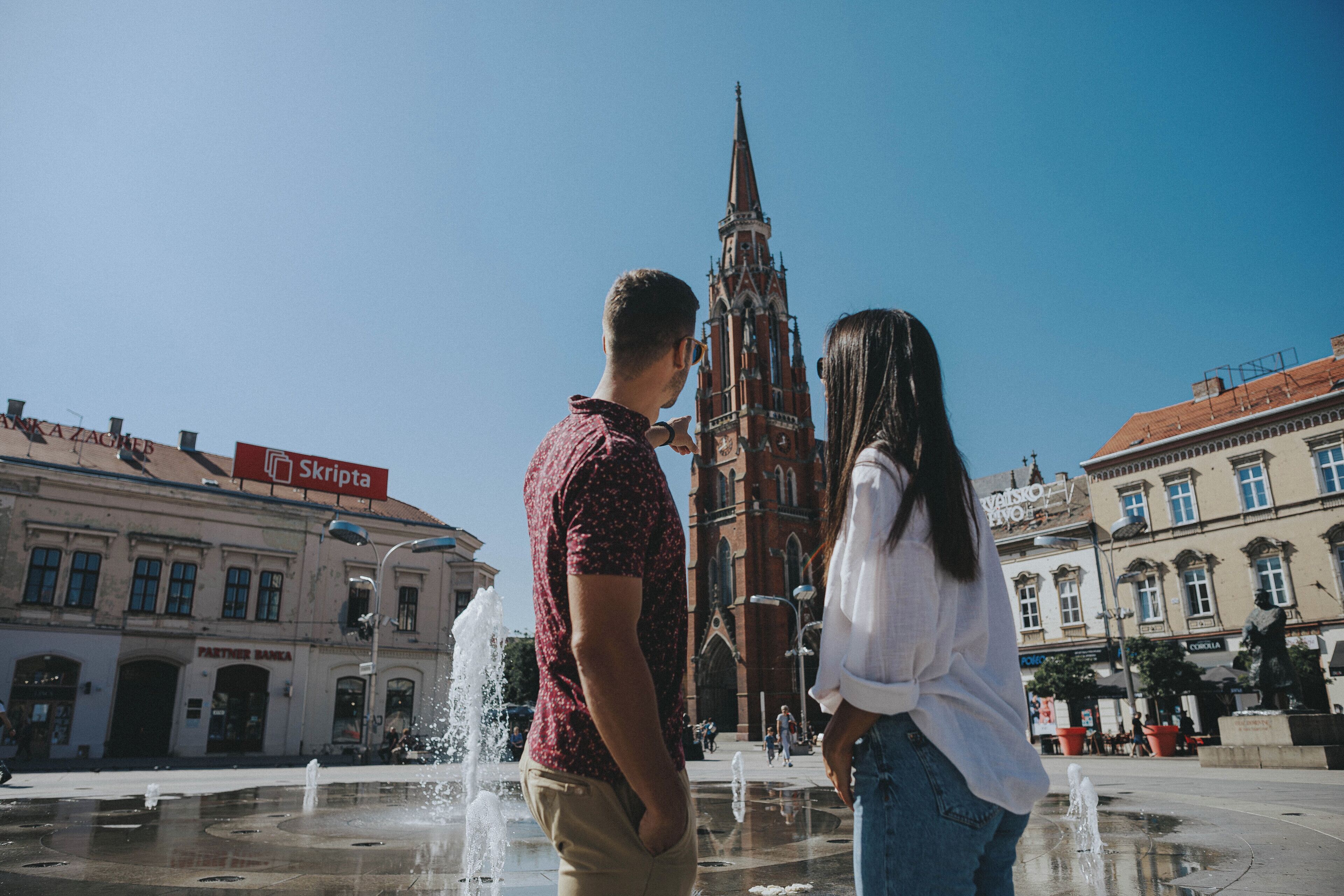 A couple standing in front of a historic church with a fountain in the foreground.