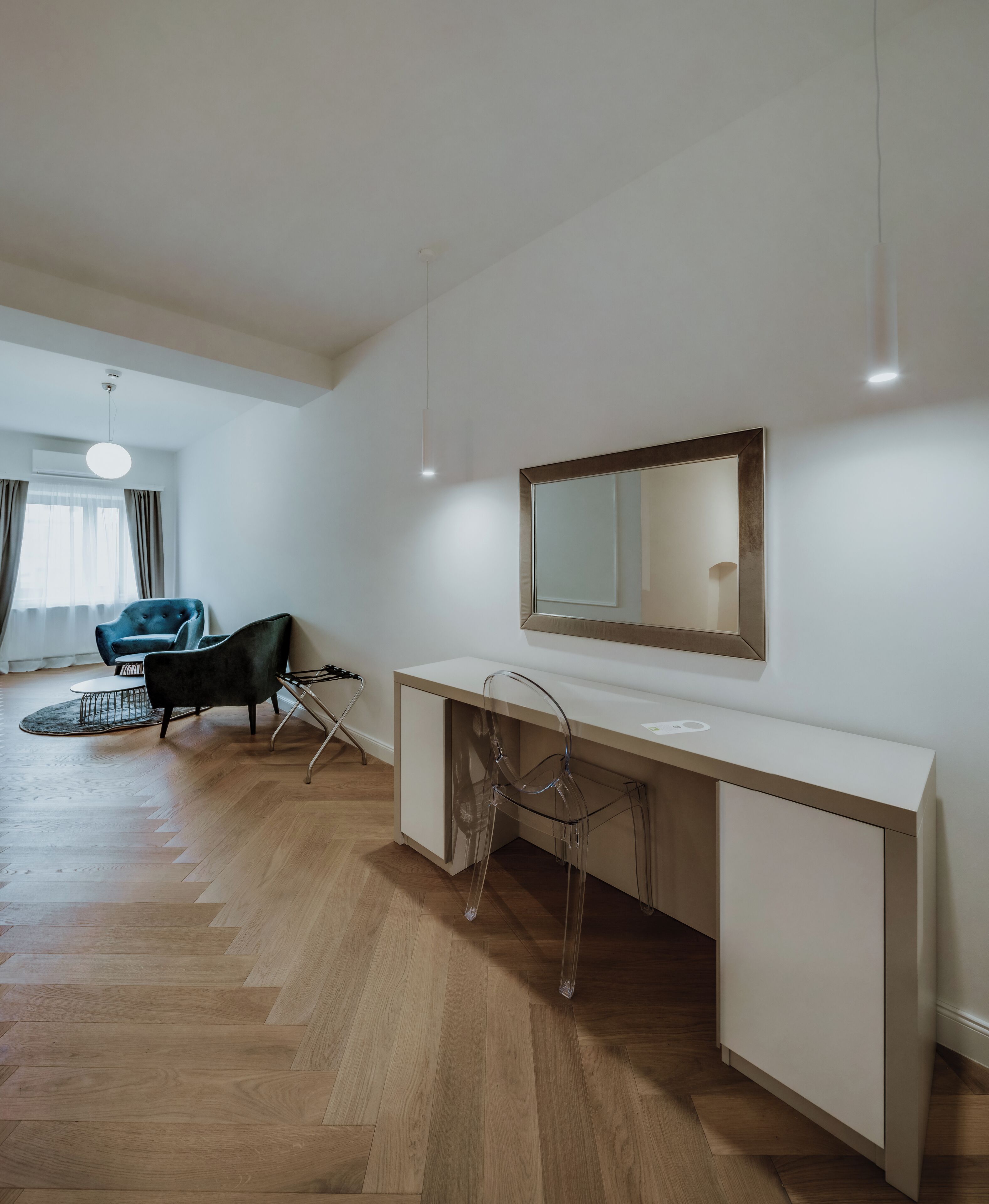 Una habitación moderna con piso de madera en patrón chevron, un escritorio blanco con espejo, una silla transparente y un sillón azul.