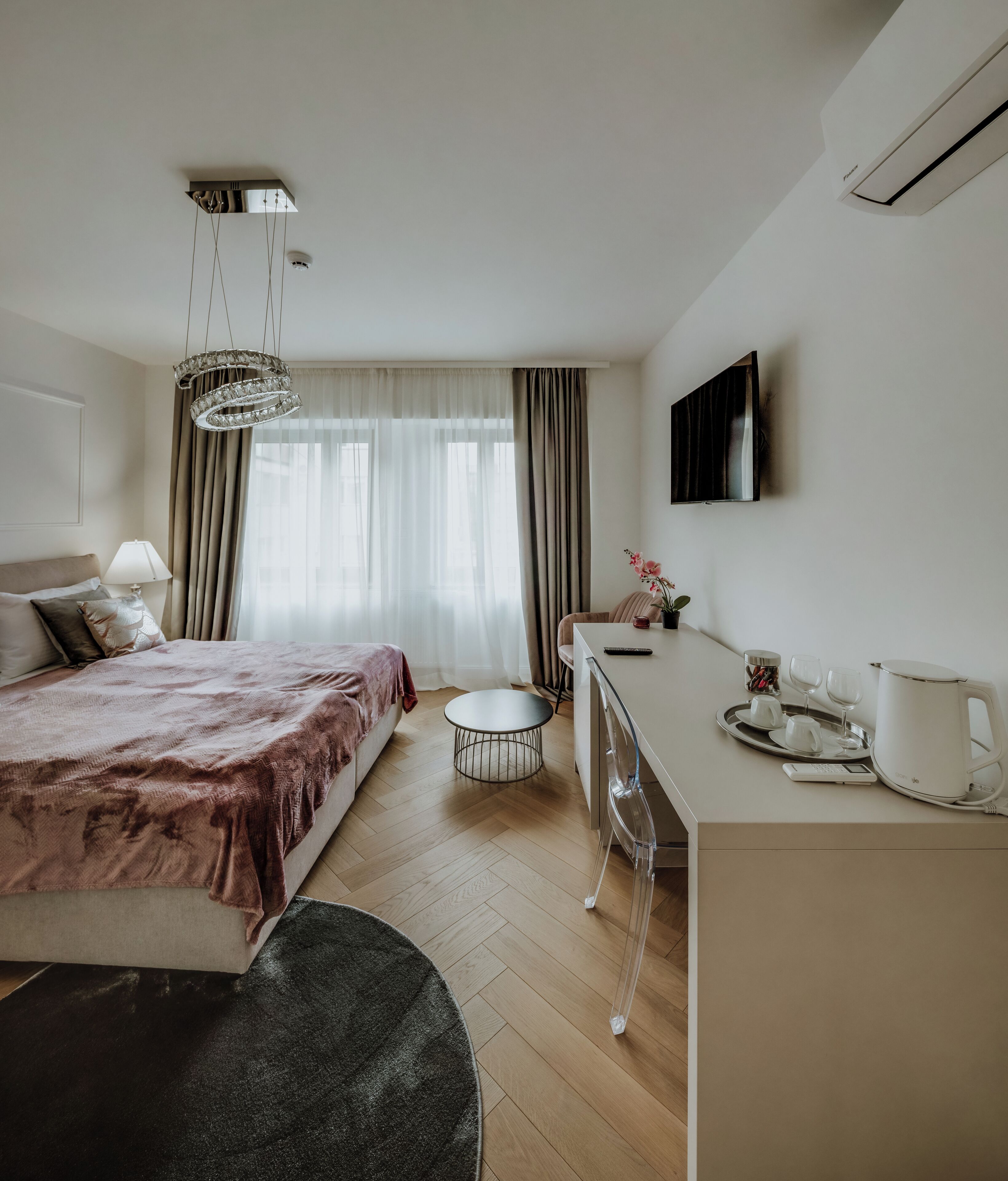 A modern hotel room with a bed, a desk, a chair, a small table, and a TV mounted on the wall.