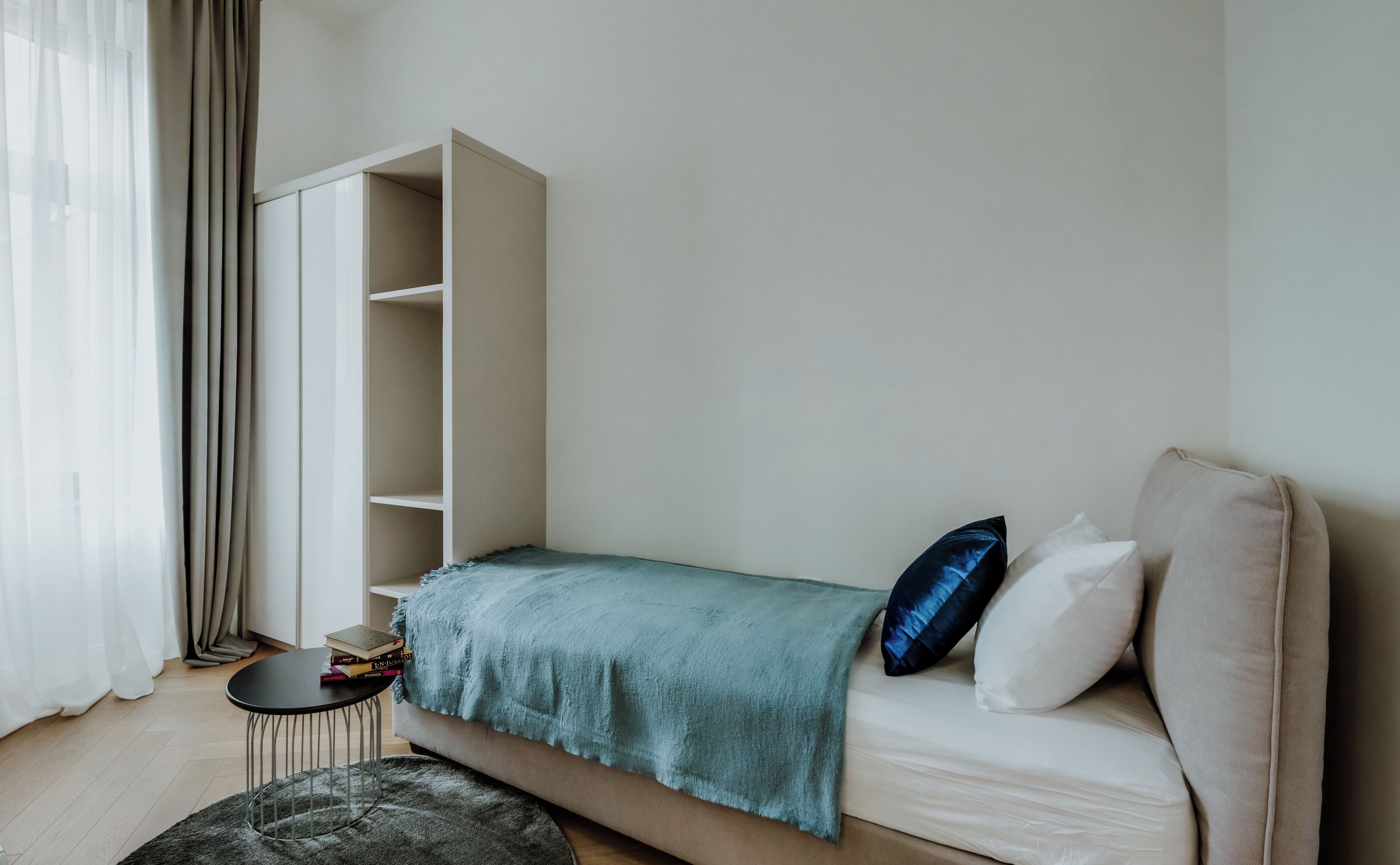 A bedroom with a bed, a side table, a bookshelf, and a window with curtains.