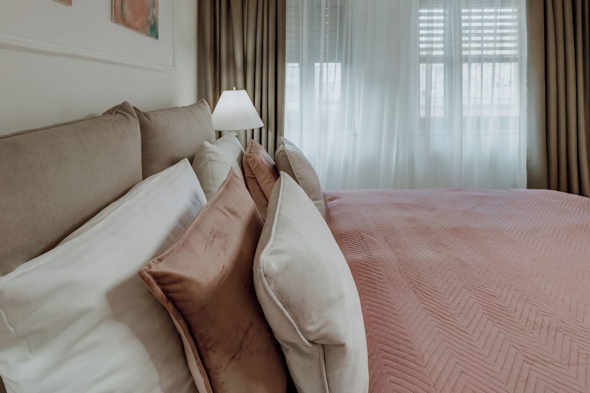 Une chambre avec un grand lit, des oreillers beiges et blancs, une lampe de chevet et des rideaux légers.
