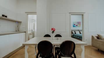 A modern kitchen with white cabinetry, a white countertop, and a black coffee maker. There is a dining area with a white table and two chairs. A beige sofa is visible in the background.