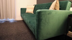 Un canapé vert avec deux coussins, une moquette foncée et un mur blanc avec une fenêtre.