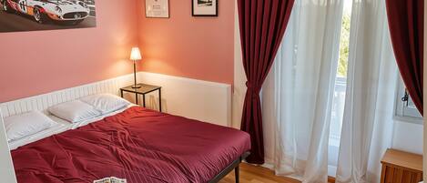 Une chambre à coucher avec un lit, une table de chevet, une lampe, un tableau encadré et une fenêtre avec des rideaux.