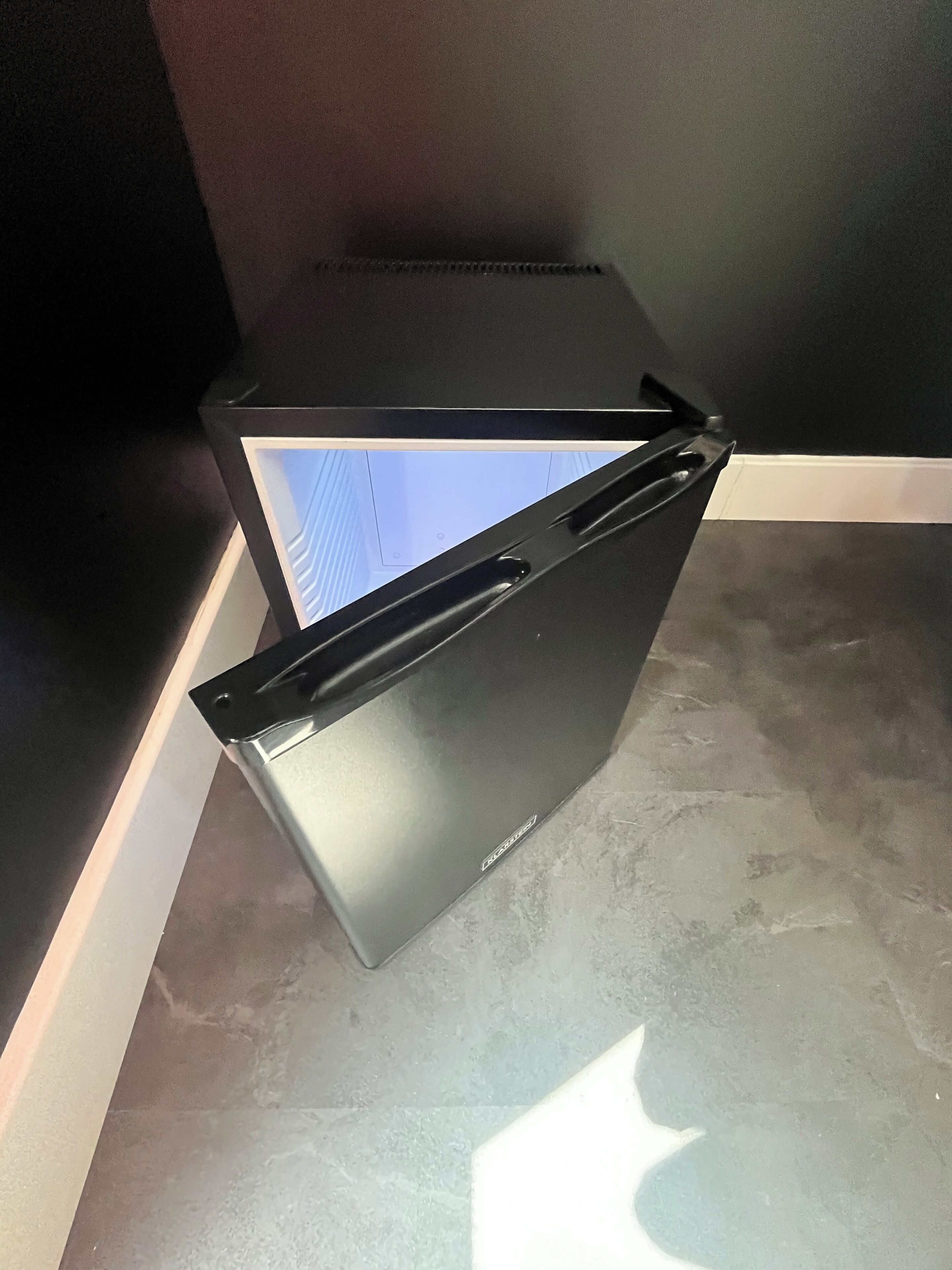 A corner of a room with a black refrigerator, a white baseboard, and a grey tiled floor.