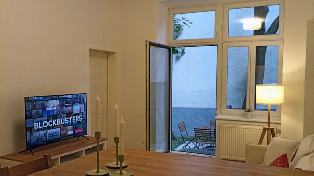 A living room with a wooden dining table, a TV displaying 'Blockbusters', and a view of the sea through a glass door. - 170m2 with private garden (Wien)