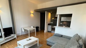 Egy modern nappali, szürke kanapéval, fehér dohányasztallal és televízióval. Egy kis konyhasarok is található benne, amelyben tűzhely és mosogató van.