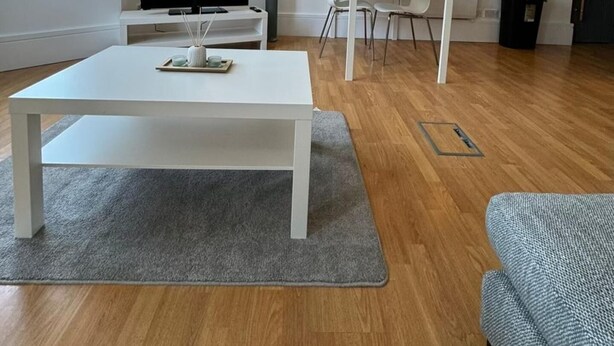 Egy modern nappali, melyben egy síkképernyős televízió, egy fehér dohányasztal és egy szürke kanapé található.