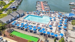 An aerial view of a resort with a large swimming pool surrounded by sun loungers and umbrellas, a dock with boats, and a river in the background. - Dolphins Chance - Waterfront Ocean Pines + Dock (Ocean Pines)