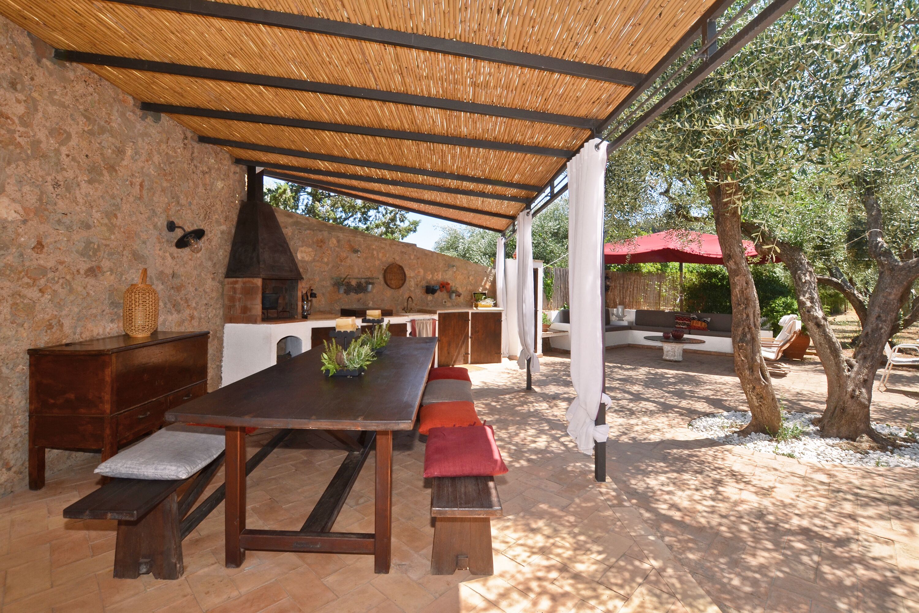 An outdoor patio with a rustic stone wall, a wooden table with benches, and a pergola overhead.