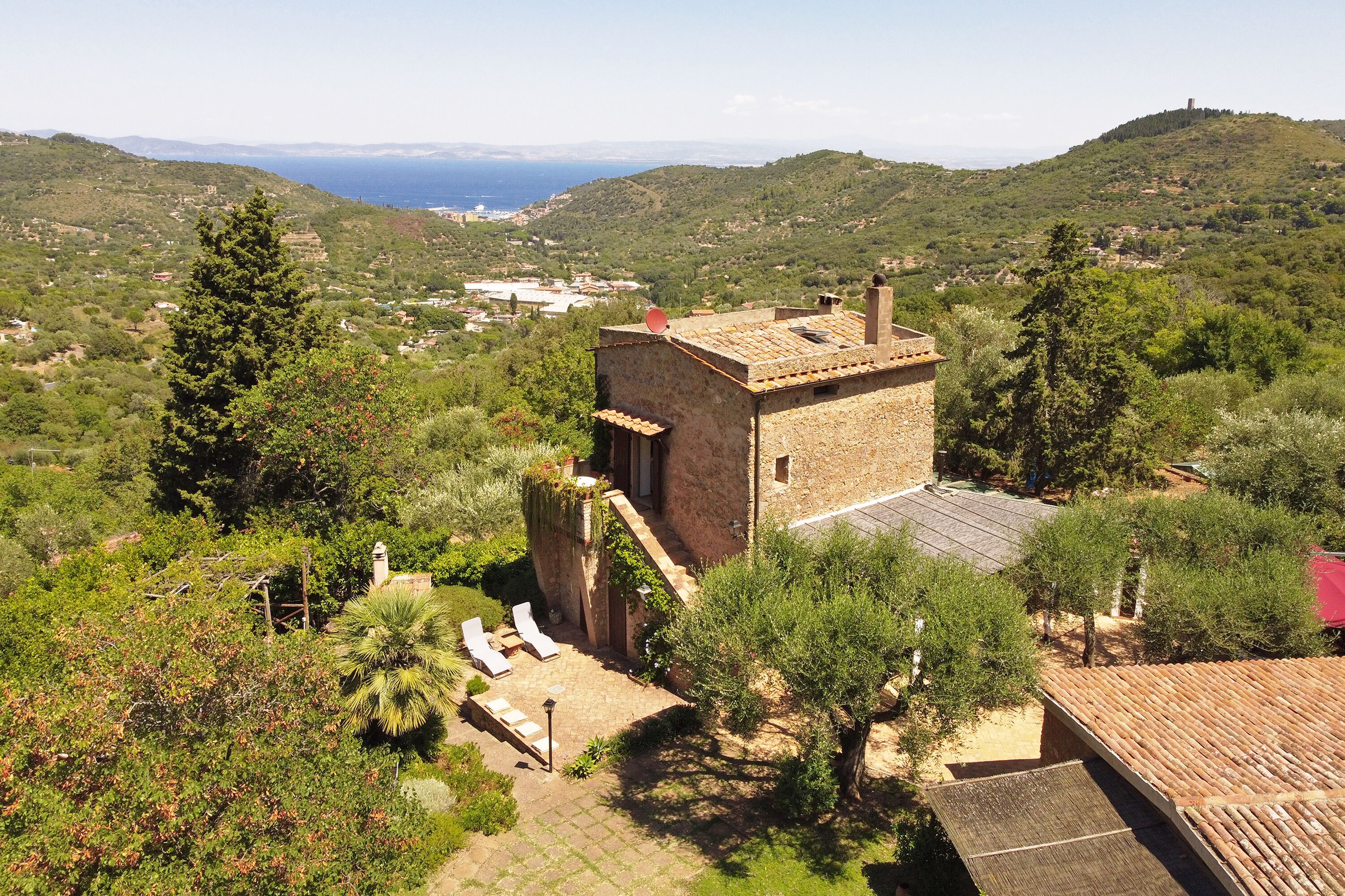Un bâtiment en pierre, doté d’un toit en tuiles, entouré d’une végétation luxuriante et offrant une vue sur la mer.