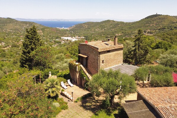 A stone building with a tiled roof, surrounded by lush greenery and a view of the sea.