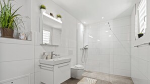 Un bagno moderno con piastrelle bianche, un lavandino, un water, una doccia e due piante in vaso sul mobile.