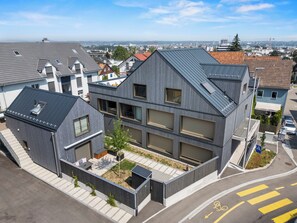 A modern residential building with a sloped roof, multiple windows, and a small outdoor patio area. - Ebenerdiges- Bodensee- Apartment mit Terrasse und Kleinem Garten (Kreuzlingen)