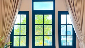 A room with three windows, each with a dark frame and a view of greenery outside. - Kim Sa Village - NovaWorld Phan Thiet (Tien Thanh)