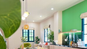 A modern dining area with a wooden table and chairs, a kitchen with a green accent wall, and a view of the outdoors. - Kim Sa Village - NovaWorld Phan Thiet (Tien Thanh)