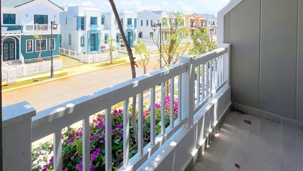 A balcony with white railings, pink flowers, and a view of a street with white buildings. - Kim Sa Village - NovaWorld Phan Thiet (Tien Thanh)