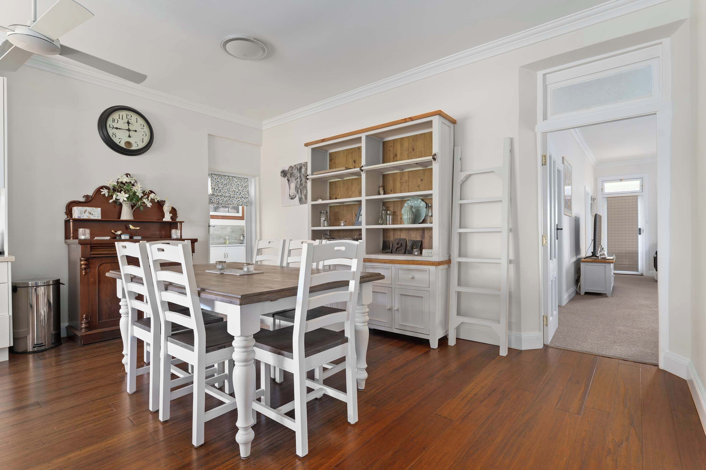 Un coin repas avec une table en bois et des chaises blanches, un meuble en bois doté de portes en verre et une horloge au mur.