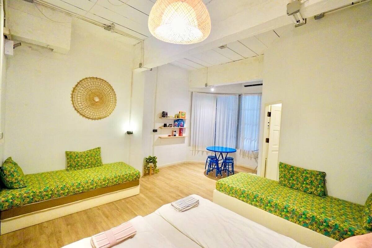 A room with two green patterned sofas, a small table with chairs, a bed, a hanging light fixture, and a wall-mounted shelf with items.