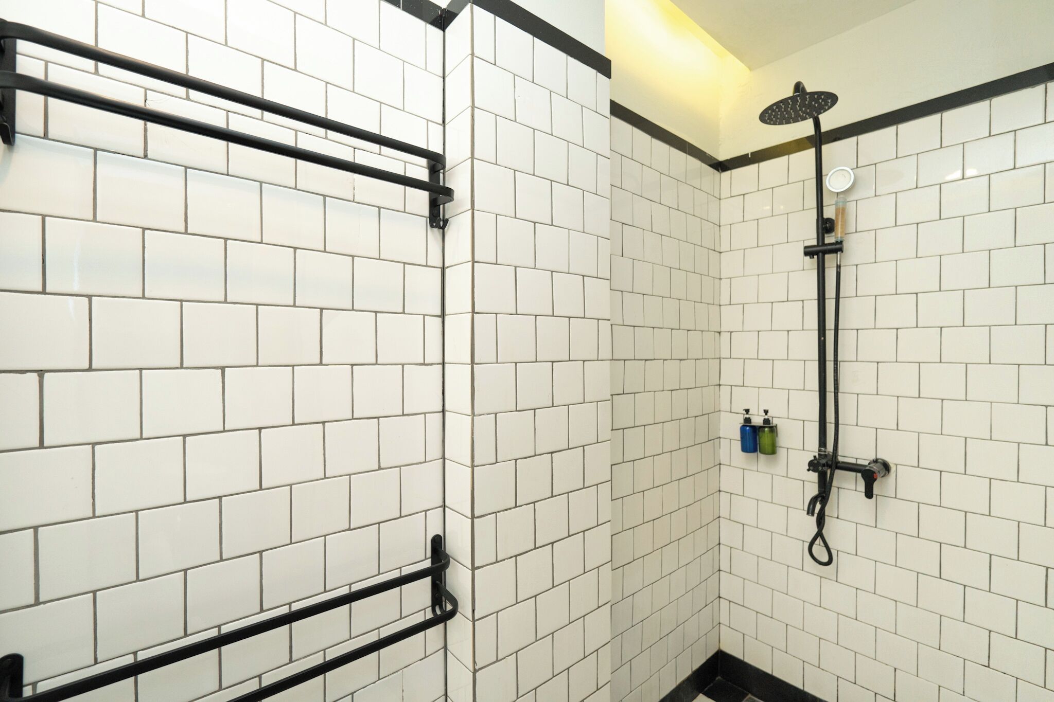 Une salle de bain avec des murs carrelés de carreaux blancs, un porte-serviettes noir, une pomme de douche et un distributeur de savon.