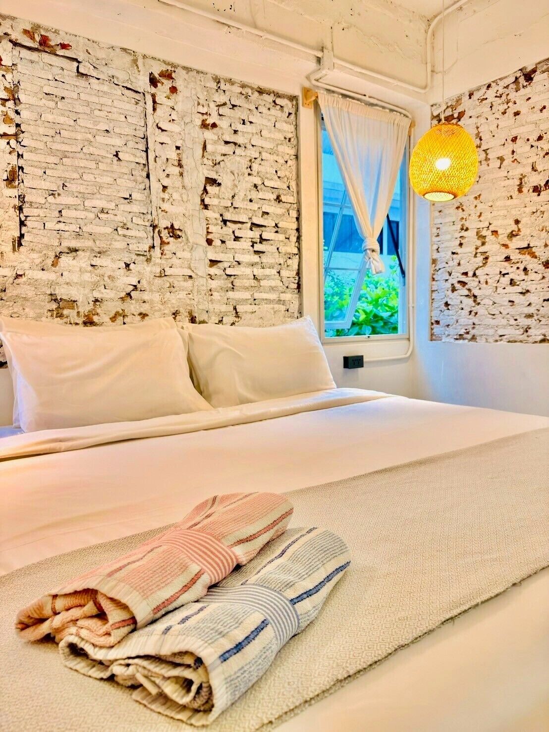 A hotel room with a bed, white curtains, a window with a view of greenery, and a textured wall.