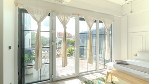 Una habitación con cama, un banco y vistas a través de grandes puertas correderas hacia un espacio exterior con plantas y un edificio al fondo.
