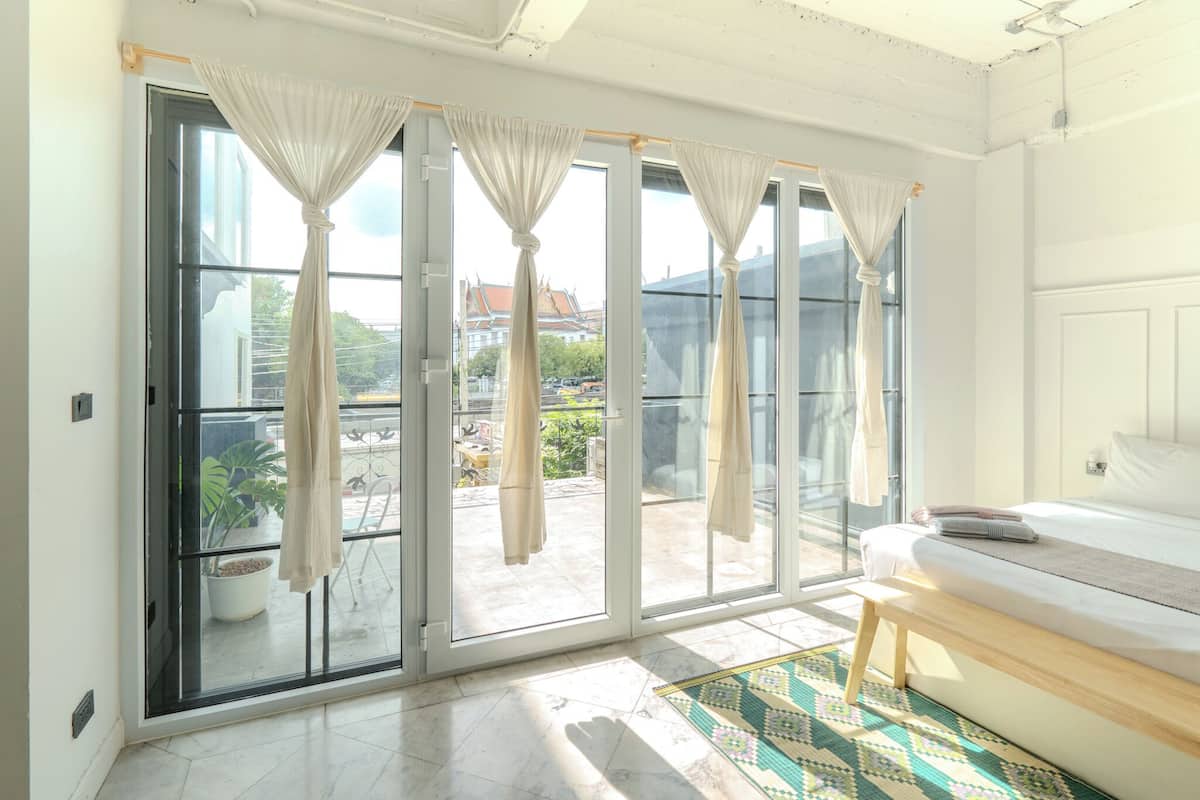 A room with a bed, a bench, and a view through large sliding glass doors to an outdoor area with plants and a building in the background.