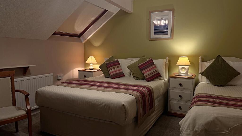 Quarto de hotel com uma cama grande, duas mesas de cabeceira com abajures, uma cadeira e um quadro na parede.