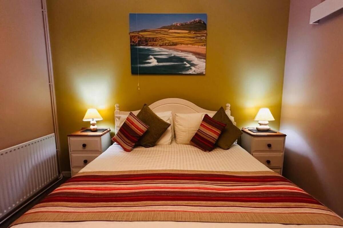 Un lit avec une couvre-lit à rayures, deux lampes de chevet et un tableau représentant un paysage côtier accroché au mur.